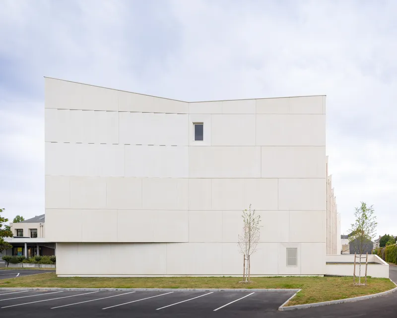 Cream-coloured precast concrete panel facade with minimal window openings flanked by young saplings and parking lot