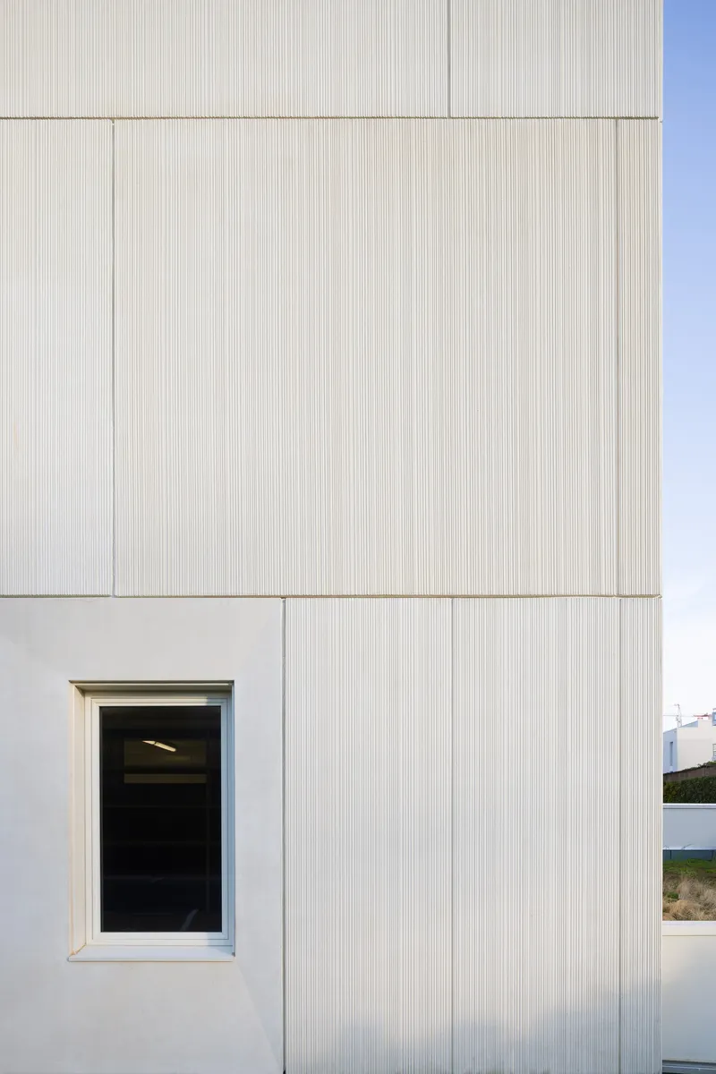 Corner of textured vertical-ribbed cladding meeting smooth panel at a single window opening below blue sky