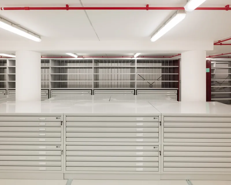 Flat file storage cabinets below wall-mounted shelving units in a white-walled archive space