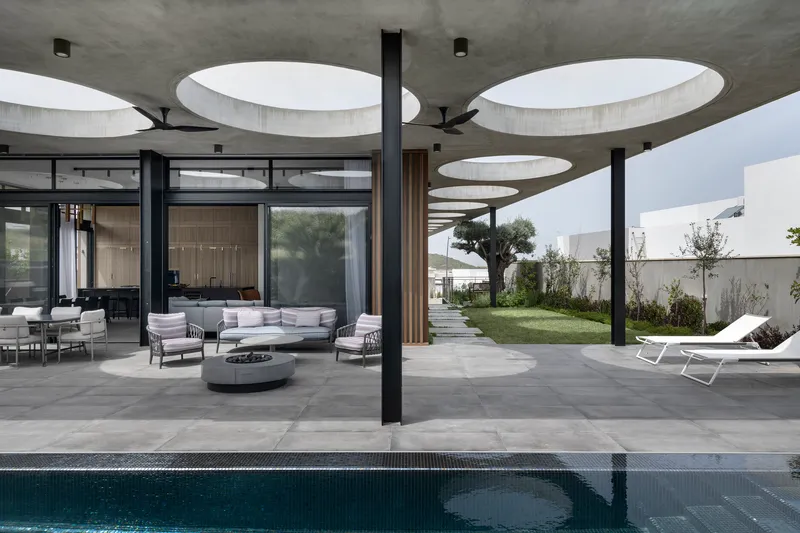 Covered outdoor terrace with circular ceiling voids and black columns overlooking a pool and garden