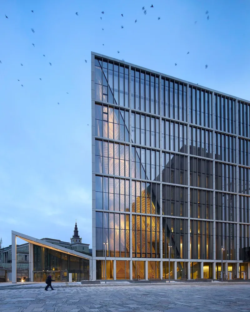 Glass facade reflecting the timber-clad atrium and surrounding buildings at dusk with birds overhead