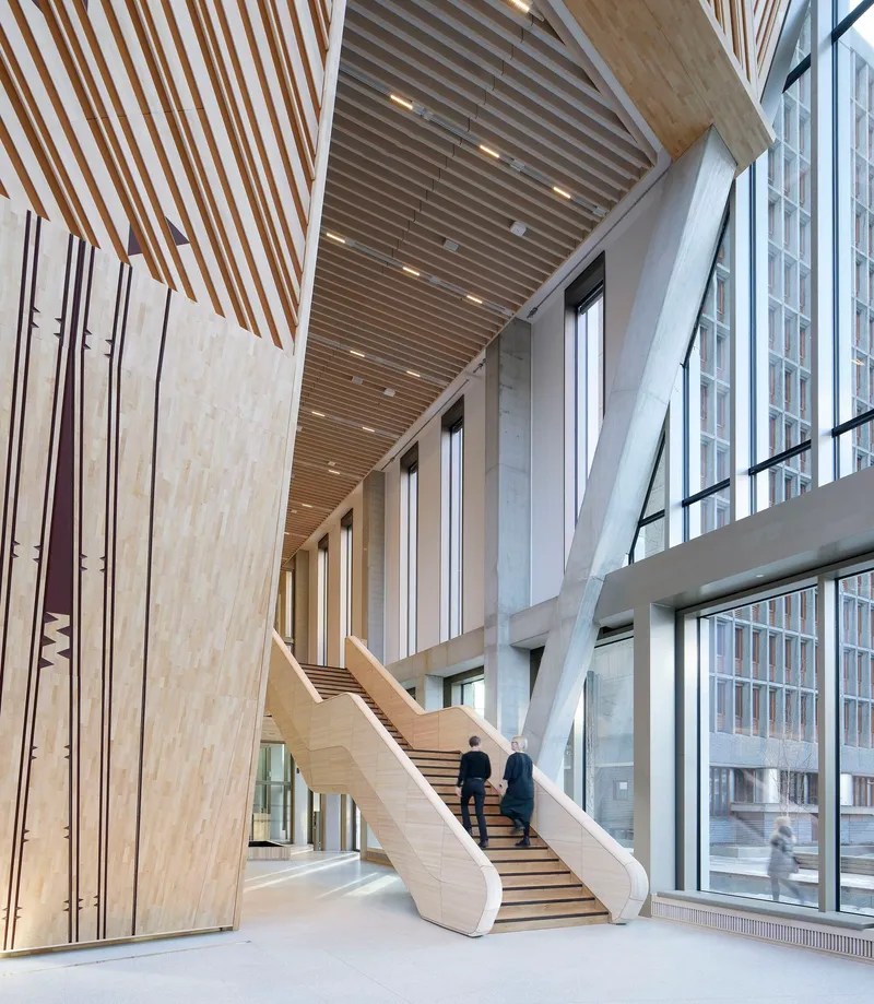 Light wood staircase ascending beneath angled structural beams and slatted ceiling with two people climbing