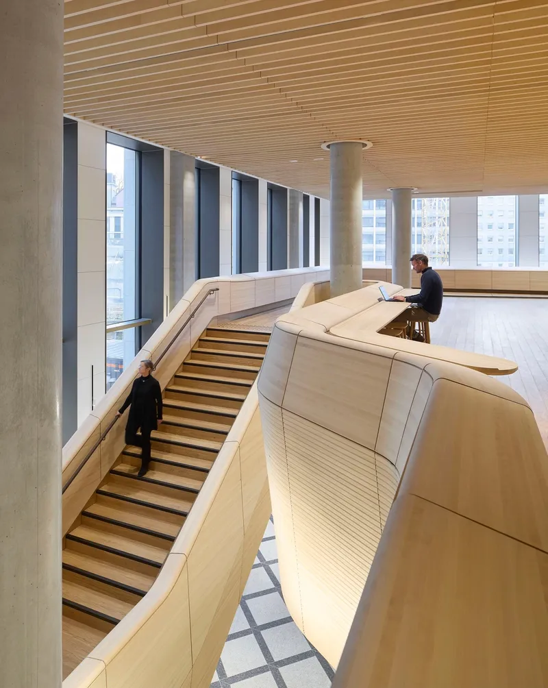 Curved bleacher seating with wood slat cladding beneath a linear wood batten ceiling as person works