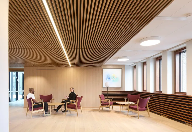 Meeting lounge with burgundy chairs beneath wood batten ceiling and circular skylights by windowed wall