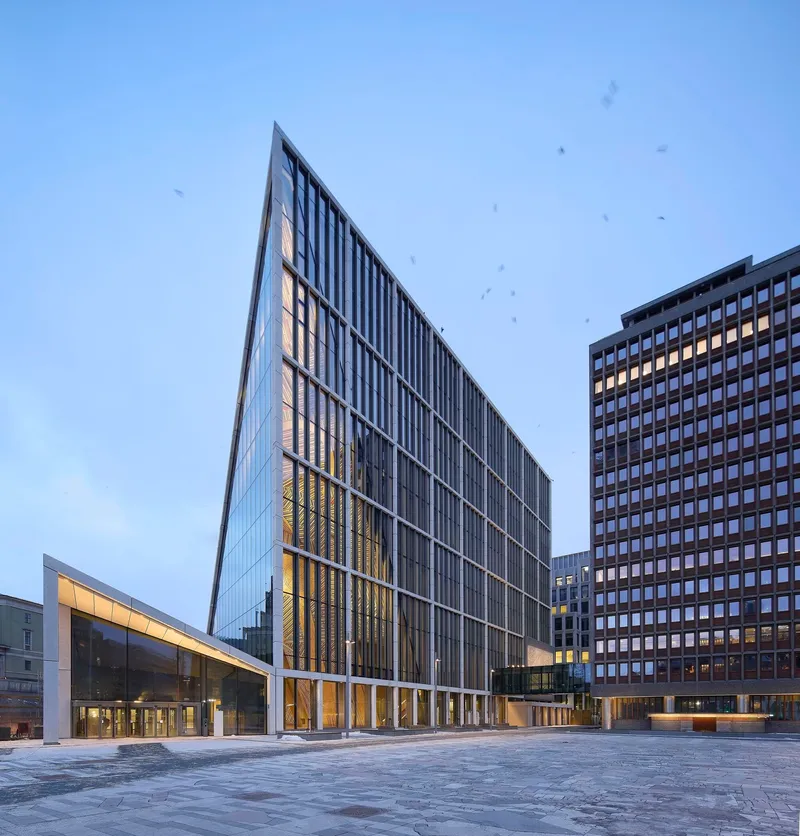 Angled glass and metal tower beside low glazed wing across paved plaza at dusk with birds overhead