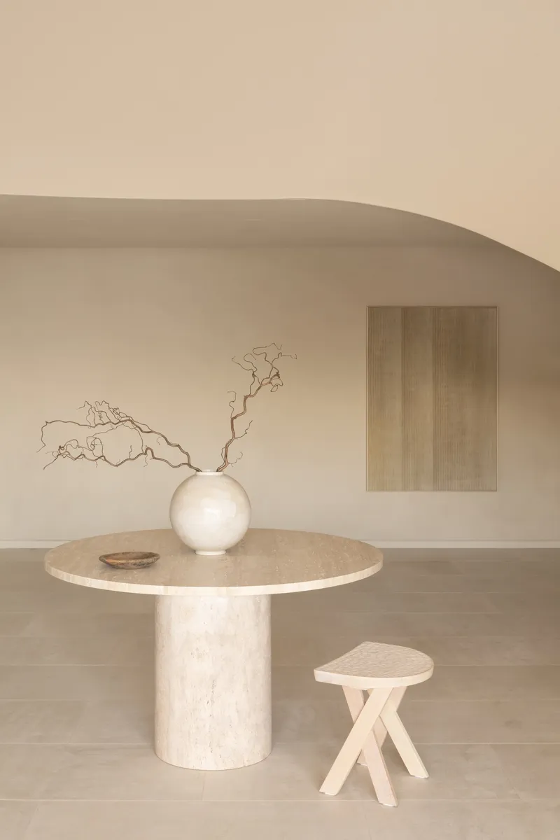 Travertine dining table with spherical vase and dried branches beneath a curved plaster ceiling