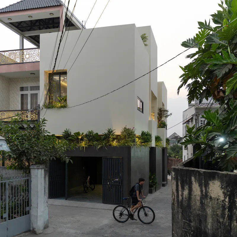 Street view of the white facade with planted base and a cyclist passing at dusk