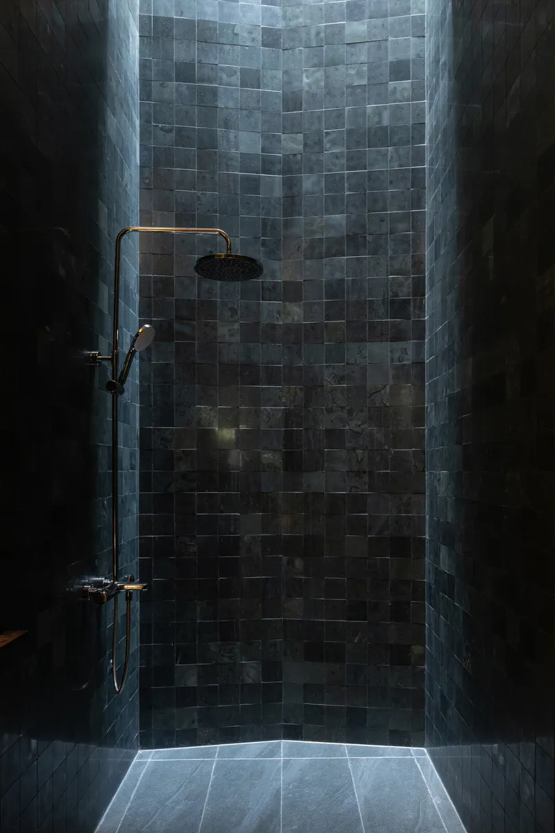 Shower enclosure with blue-grey glazed tile wall, brass fixtures, and backlit floor slit
