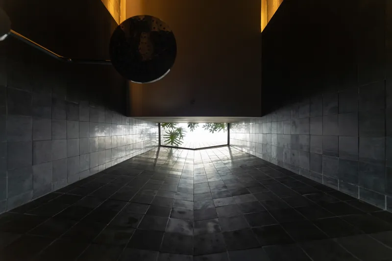 Covered entrance passage with dark stone floor and slatted walls leading to a sunlit courtyard with greenery
