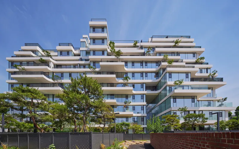 Terraced residential building with staggered white balconies and floor-to-ceiling glass behind mature pines