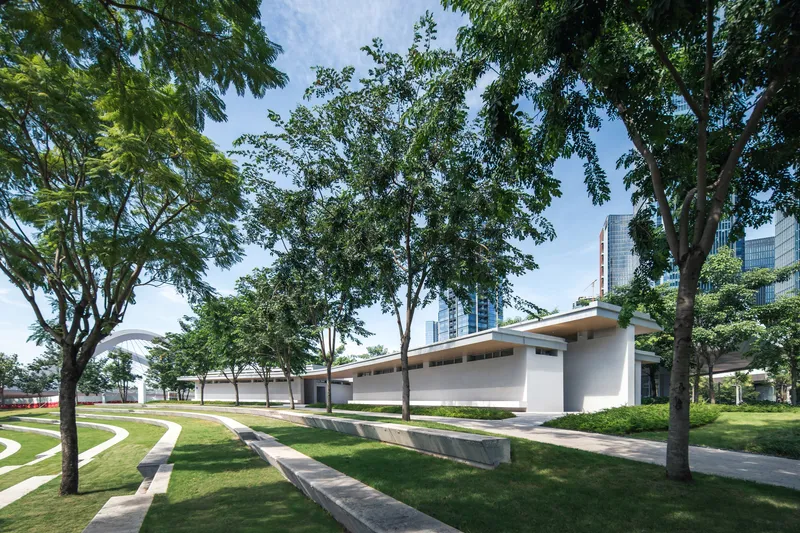 Low-slung roof volume extending through a park with curved pathways and mature canopy trees