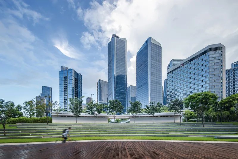 Terraced lawn with timber deck and stone seating steps facing a low horizontal pavilion and glass towers