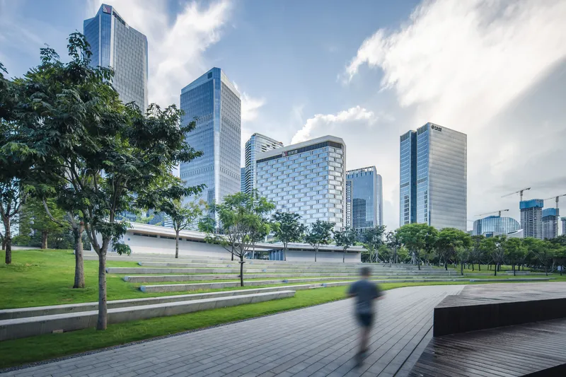 Curving paved plaza with terraced lawn and a pedestrian walking past scattered trees in daylight