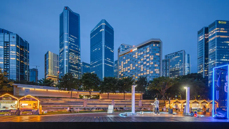 Low white pavilion with illuminated market stalls and timber deck at dusk beneath a tower skyline