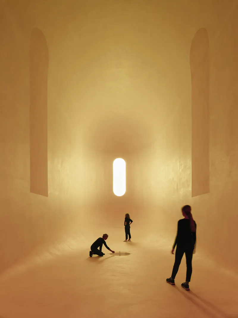Golden-lit vaulted interior with tall vertical niches and visitors gathered beneath a single capsule-shaped window