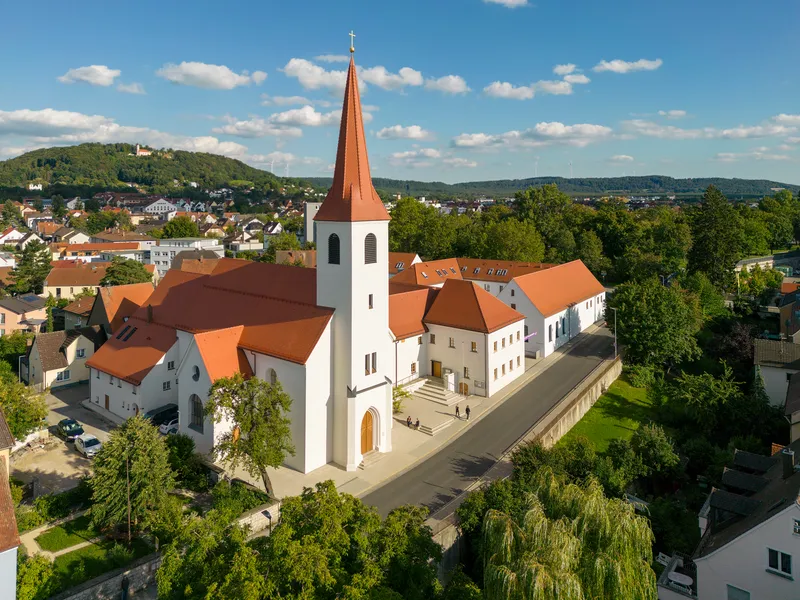 Aerial view of a white-walled church with terracotta roof and tall spire surrounded by summer greenery