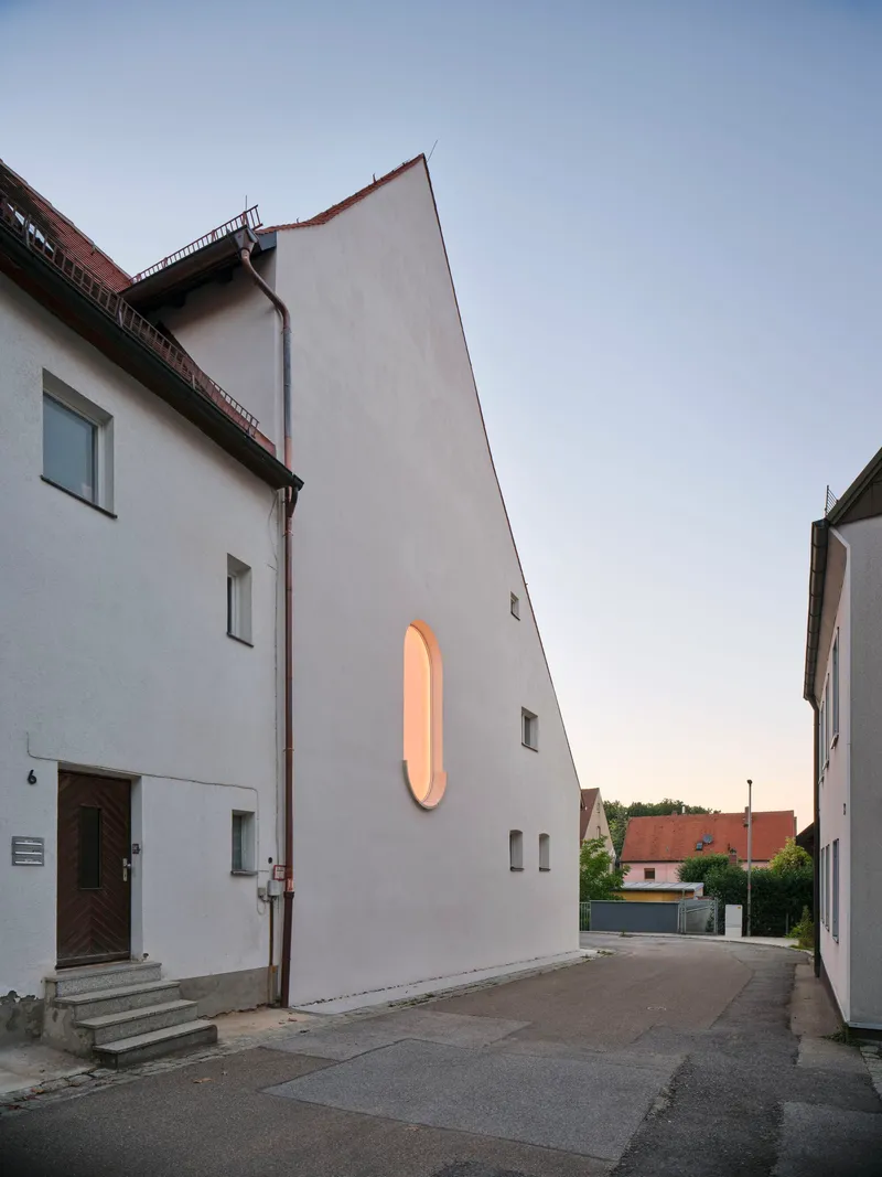 White rendered facade with an illuminated oval window opening facing a quiet street at dusk