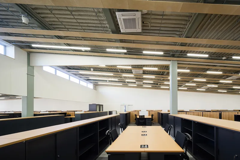 Open office workspace with exposed corrugated metal ceiling, linear lighting, and rows of black storage units