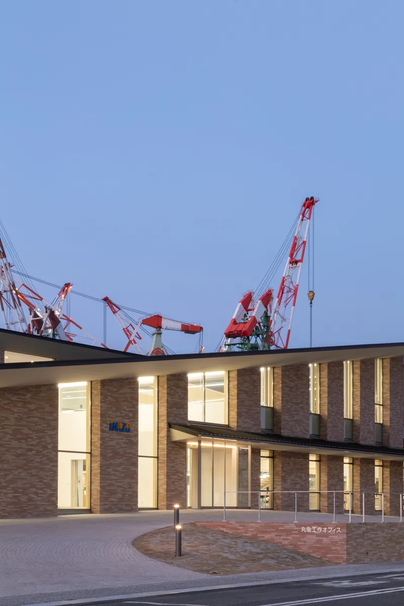 Two-story brick facade with floor-to-ceiling glazing illuminated at dusk beside the curved paved forecourt