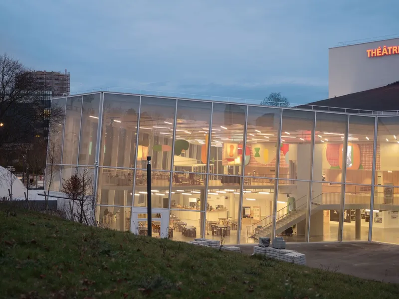 Glass-walled facade illuminated at dusk with young trees on the sloped forecourt