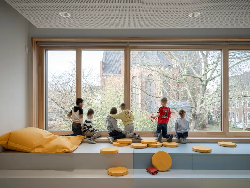 Window bench with children seated facing bare branches and brick buildings outside