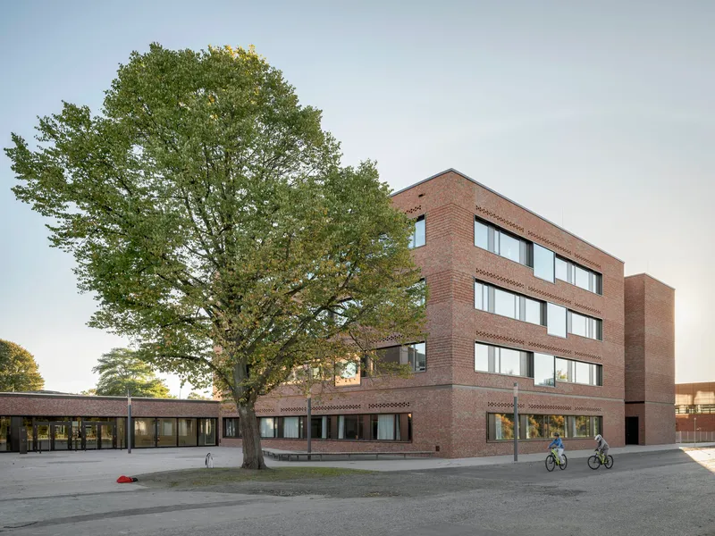 Four-storey brick building with perforated bands and a mature tree in the forecourt