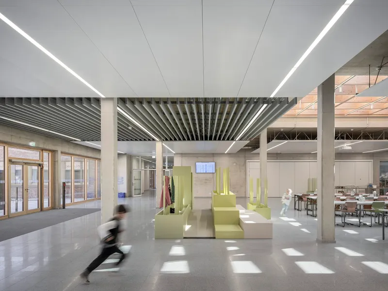 Double-height foyer with linear ceiling baffles, concrete columns and a person running through sunlight
