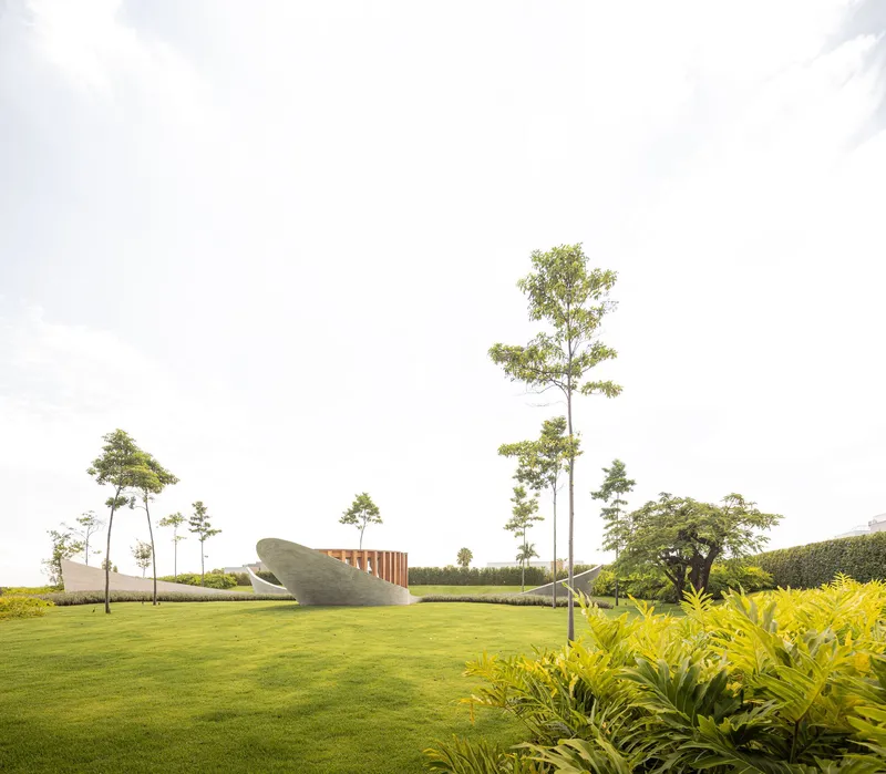Curved concrete volumes with vertical timber fins set in a manicured lawn with young trees and dense planting