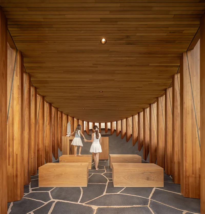 Timber-lined corridor with vertical slat walls and grey flagstone paving as two figures descend the passage