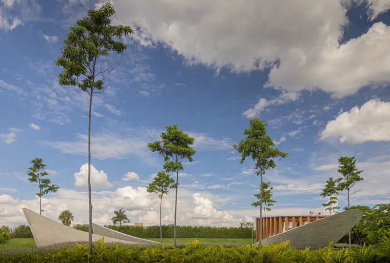 Curved concrete roof forms rising from green lawn among tall trees under scattered clouds
