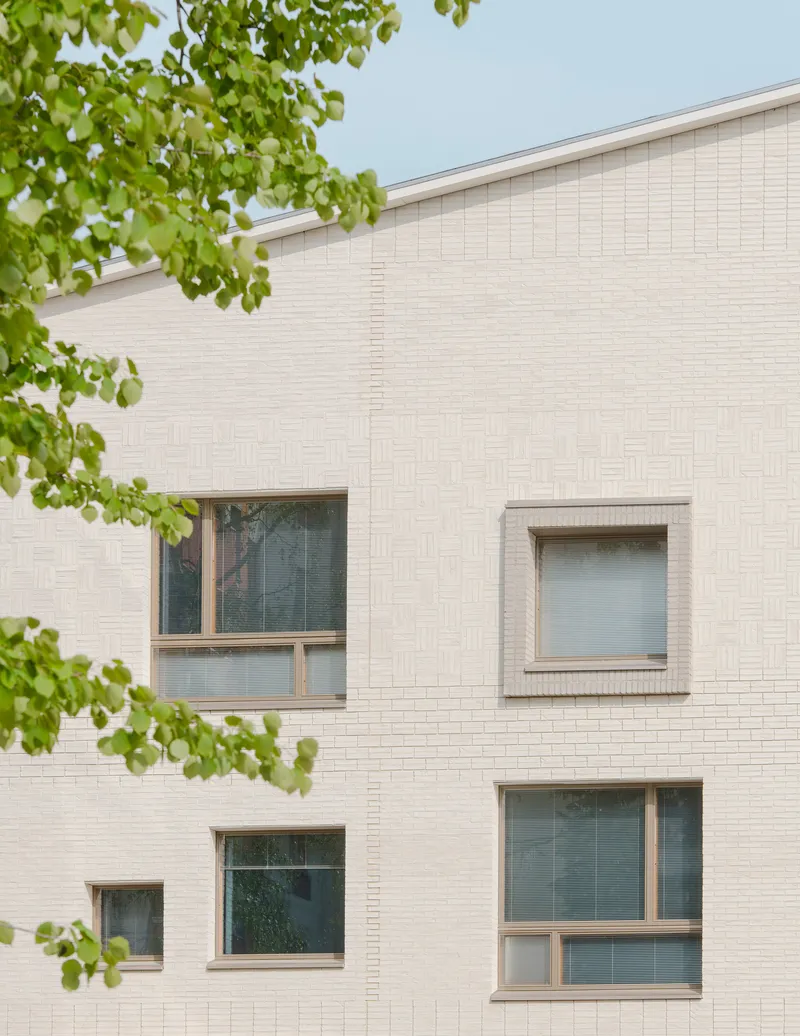 Pale brick facade with scattered windows framed by green tree branches under overcast sky