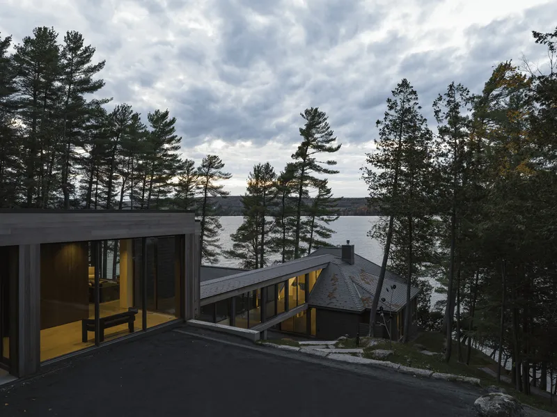 Exterior view of dark-clad volumes among pines overlooking a lake at dusk