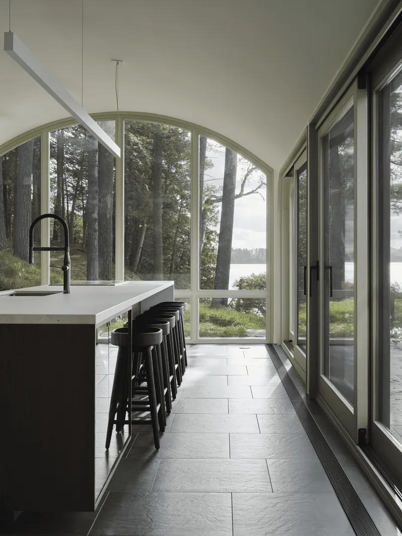 Kitchen island with black stools beneath arched windows framing pine trees and water beyond