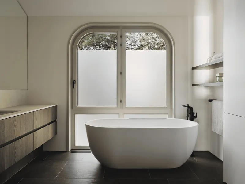 Freestanding bathtub below arched window with frosted lower panes and tree views