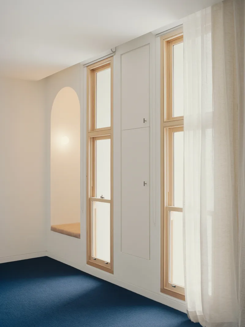 Interior wall with two arched alcoves featuring timber-framed benches and tall vertical windows with sheer curtains
