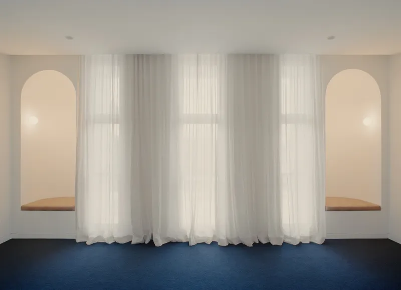 Symmetrical wall with twin arched niches and timber window seats behind floor-length sheer curtains over blue carpet