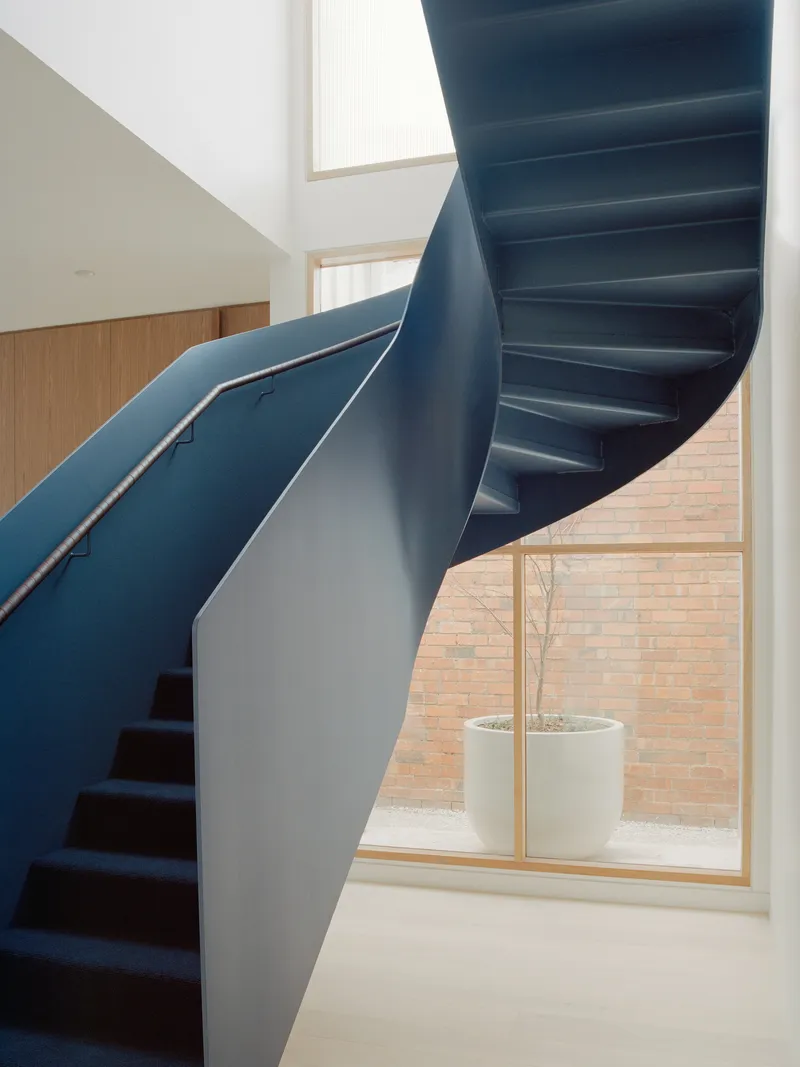 Sculptural blue steel staircase twisting upward past a tall window with planted courtyard beyond