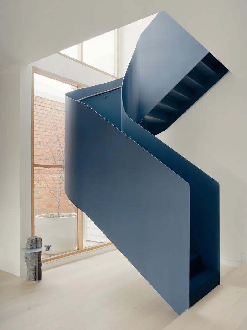 Blue ribbon staircase spiraling through void with view to brick wall and planted tree