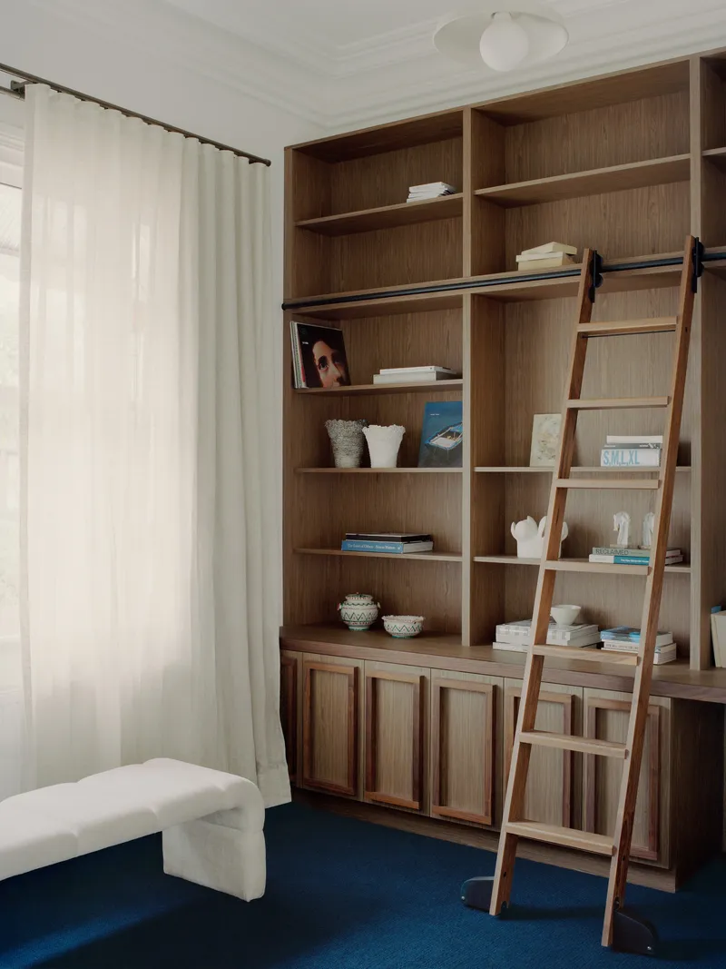 Built-in timber shelving unit with rolling ladder against blue carpeted floor and white curtains