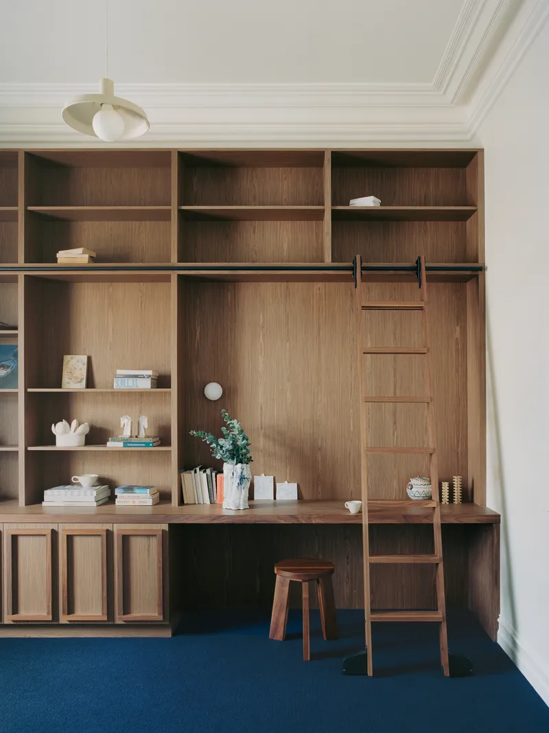 Timber desk unit integrated into shelving wall with rolling ladder and wooden stool on blue carpet