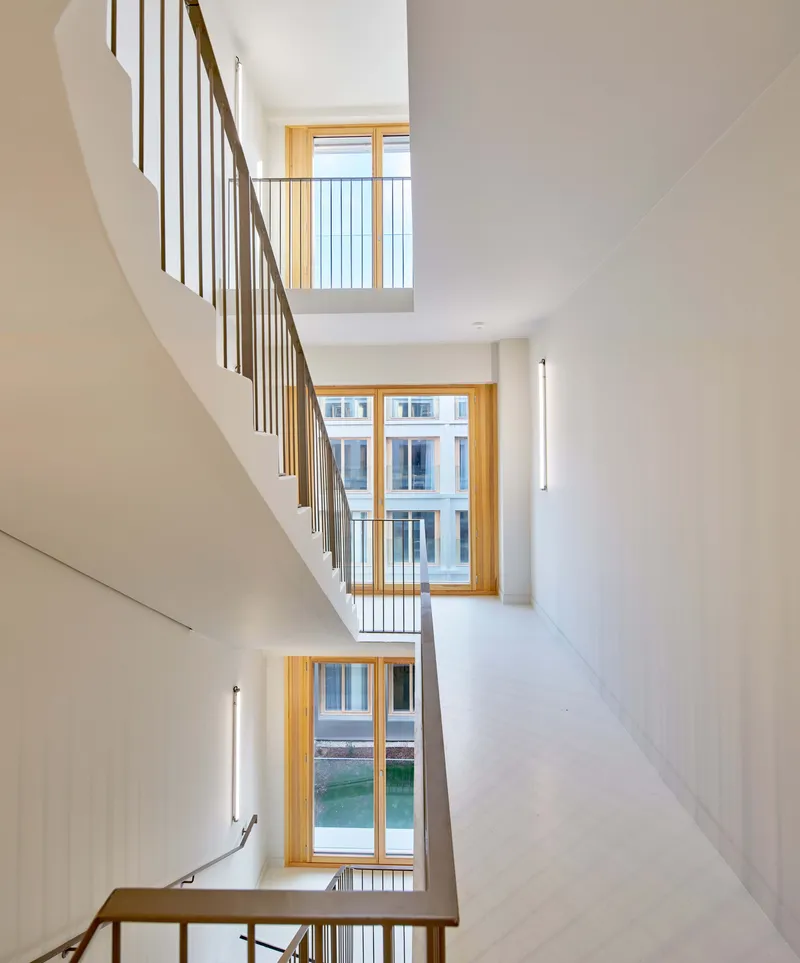 Multi-story stairwell with timber handrails and white walls illuminated by timber-framed windows at each landing
