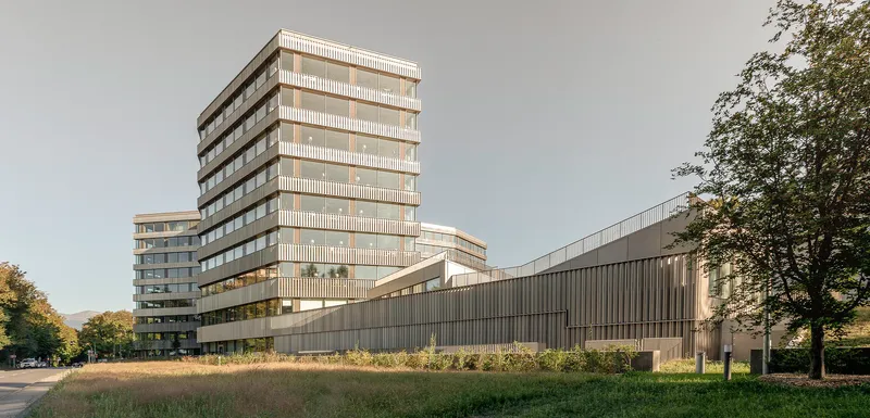 Facade composition with vertical louvered tower and horizontal ribbed base across a wildflower meadow