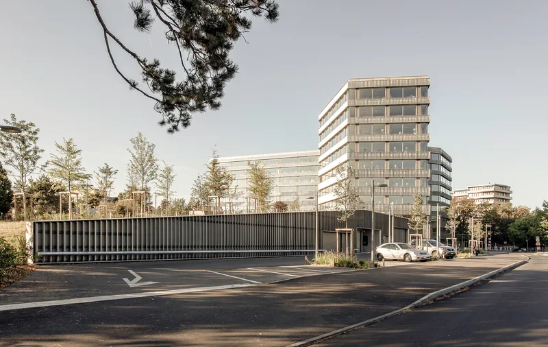 Low-rise ribbed volume with parking area and young trees under an overcast sky