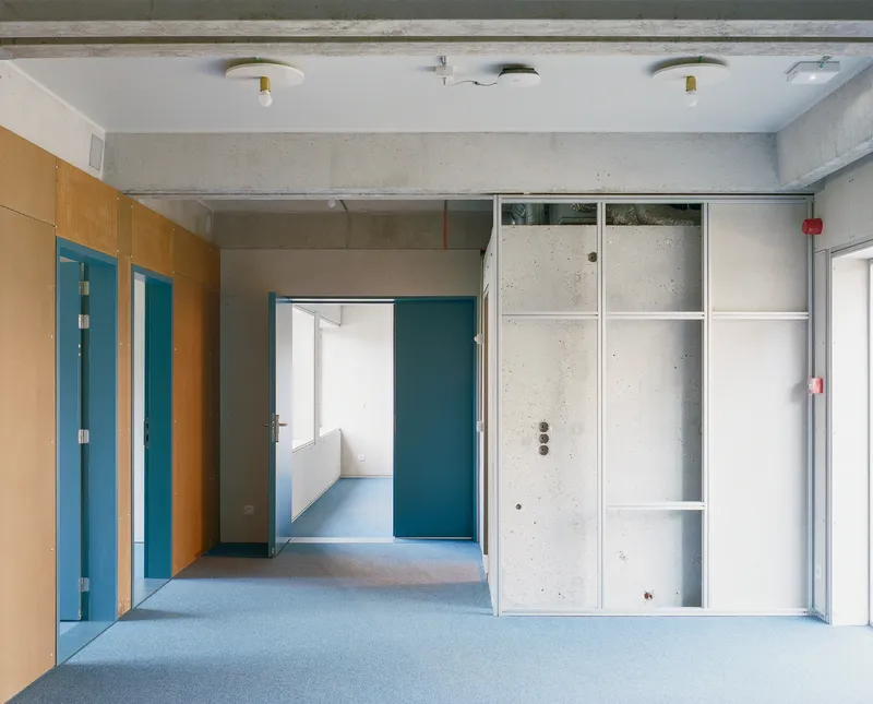 Corridor with exposed concrete ceiling, blue doors, orange plywood panels and white metal storage partitions