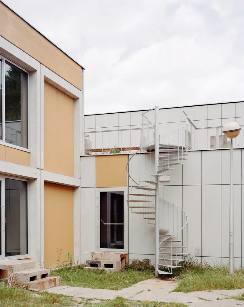 Exterior white spiral stair against white tile cladding with grassy courtyard and orange panel volumes