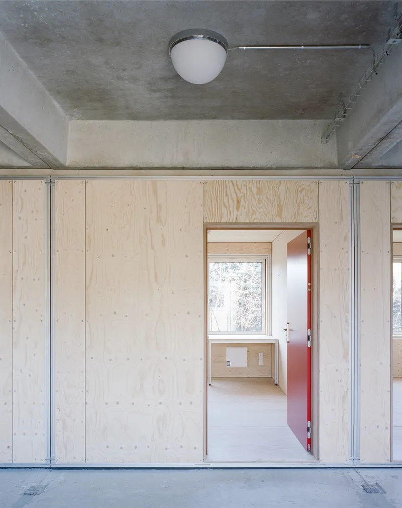 Light plywood wall with salmon-colored door opening to room with exposed concrete ceiling and steel channels