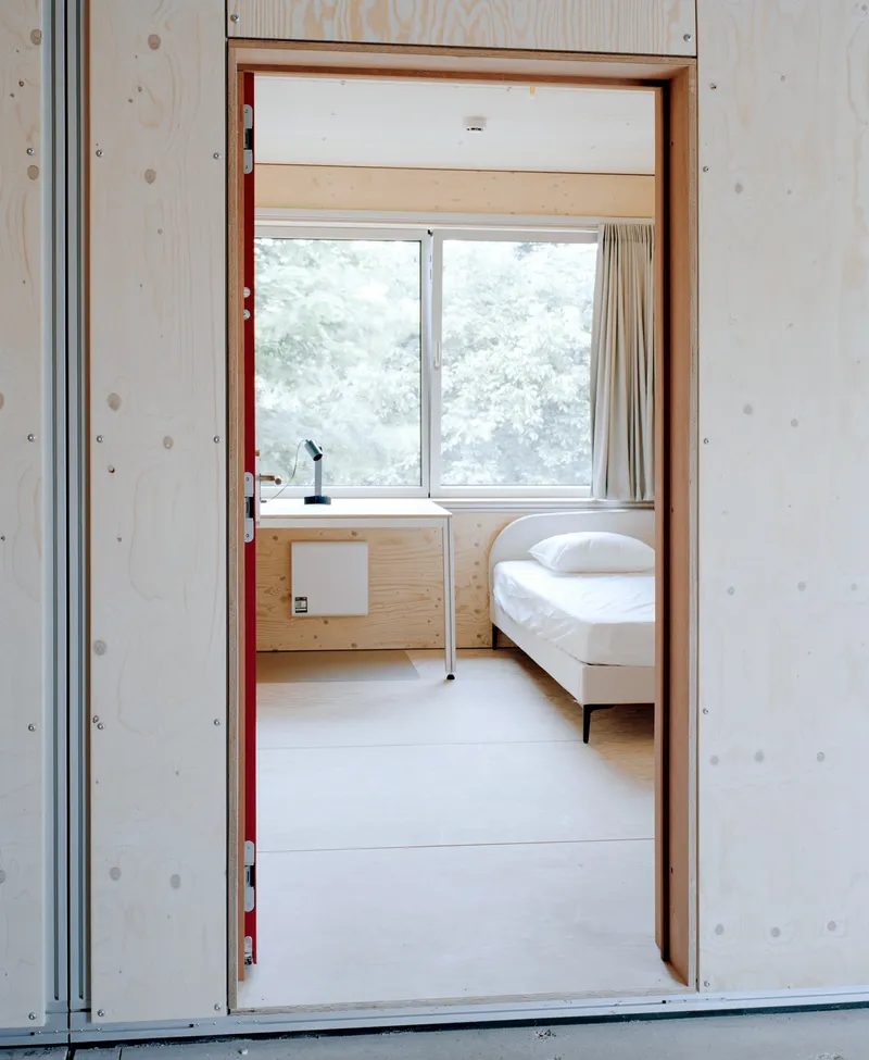 View through plywood-framed doorway into bedroom with single bed and window overlooking trees