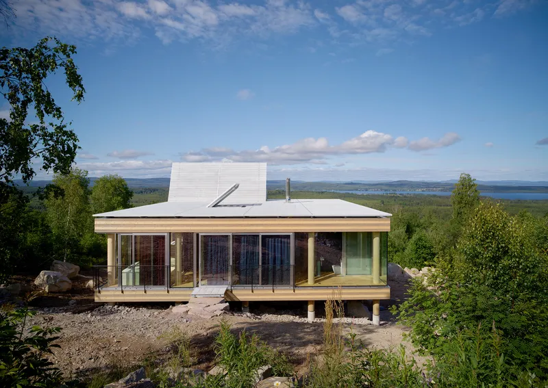 Elevated glass pavilion with white metal roof overlooking forested landscape and distant water under blue sky
