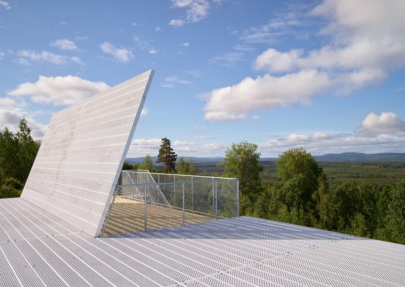 Angled white metal roof panel with chain-link fenced terrace rising above forested valley and mountains