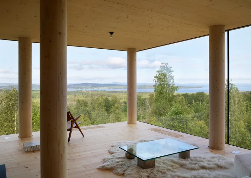 Corner room with cylindrical timber columns and plywood floor opening to lake and forest panorama
